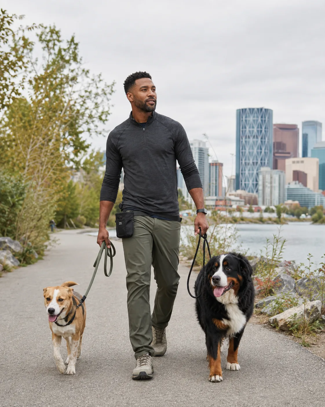 A Calgary dog walker on the river pathway with two dogs, downtown towers behind him.